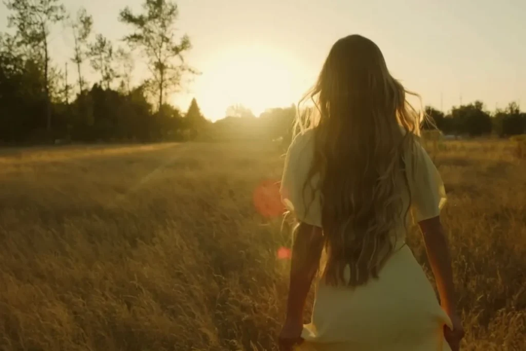 Perempuan berjalan di padang rumput pada pagi hari dengan cahaya matahari hangat, melambangkan proses mencintai diri secara perlahan.